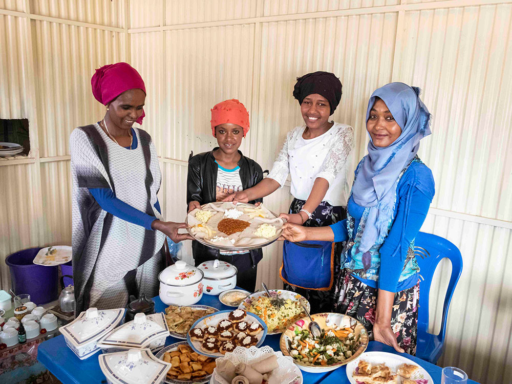 Four young Ethiopian women stand around a table full of food and present a plate of sourdough flatbread and cream cheese.