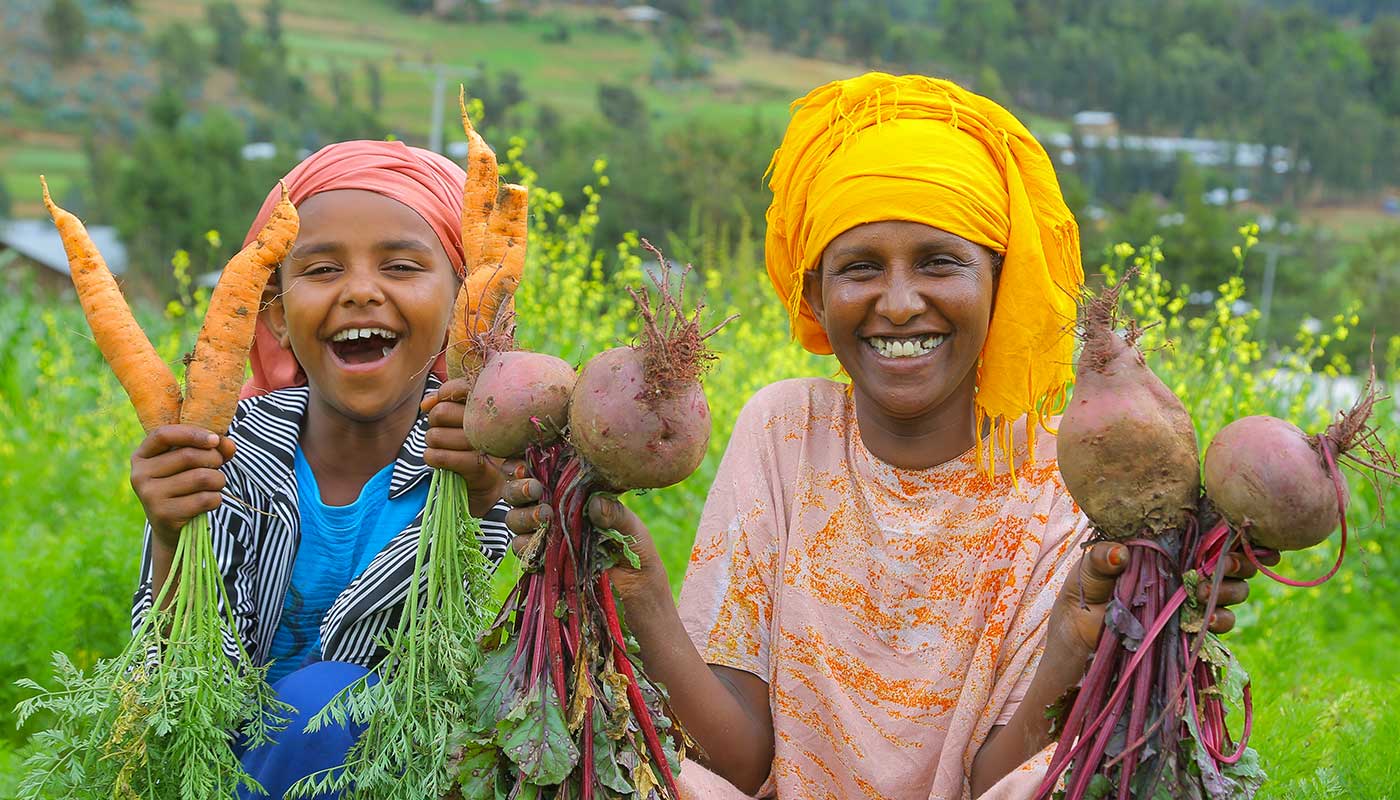 A mother and her young daughter are sitting in a field, the mother is holding red beets in her hands, the daugter carrots; both are smiling