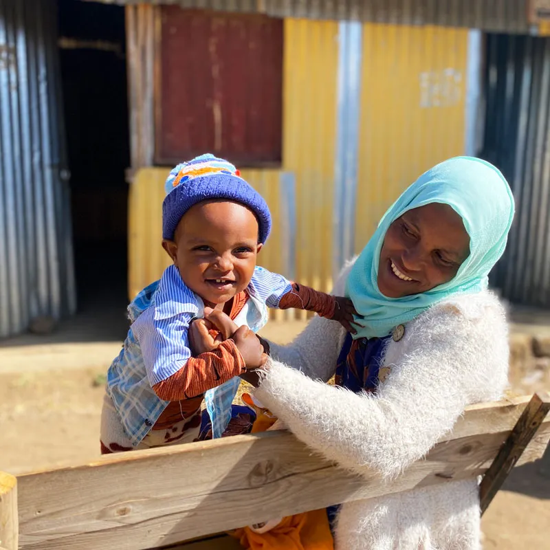 A woman in a light blue headscarf and white sweater holds a smiling toddler.