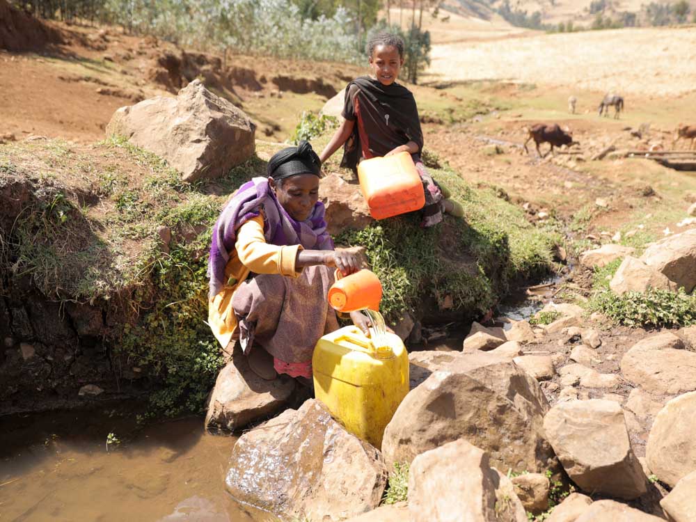 Frau und Tocher in Äthiopien holen Wasser mit gelben Kanister aus dreckigem Rinnsal